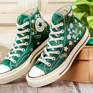 Puede incluir: Un par de zapatillas altas de lona verde con cordones y suelas blancas. Las zapatillas presentan dise&ntilde;os florales bordados en rosa, azul y blanco. El logotipo de Converse es visible en el lateral.