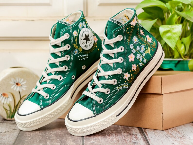 Puede incluir: Un par de zapatillas altas de lona verde con cordones y suelas blancas. Las zapatillas presentan dise&ntilde;os florales bordados en rosa, azul y blanco. El logotipo de Converse es visible en el lateral.