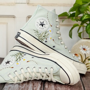 May include: A pair of light blue high-top sneakers with white soles and white laces. The sneakers are embroidered with flowers and bees. The Converse All Star logo is visible on the side of the sneakers.