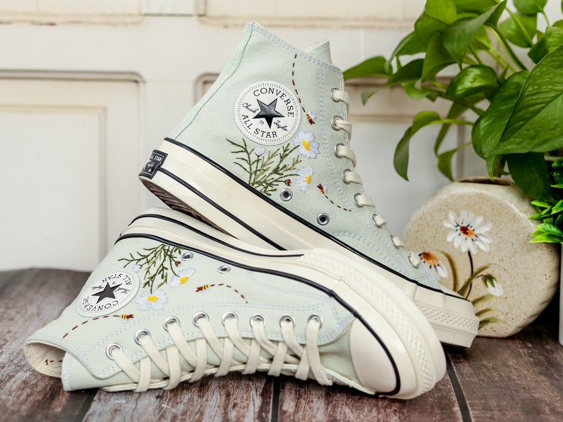 May include: A pair of light blue high-top sneakers with white soles and white laces. The sneakers are embroidered with flowers and bees. The Converse All Star logo is visible on the side of the sneakers.