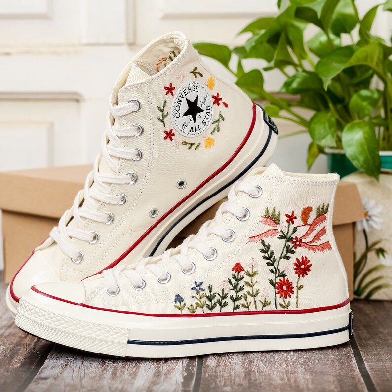 May include: A pair of white high-top canvas trainers with embroidered floral designs. The trainers have a black star logo with the text "Converse All Star" and a red stripe around the sole.