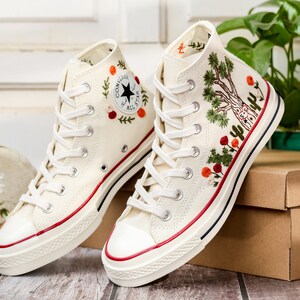 May include: A pair of white high-top canvas sneakers with red and green embroidered floral designs. The sneakers have a white rubber sole and a red stripe around the top.