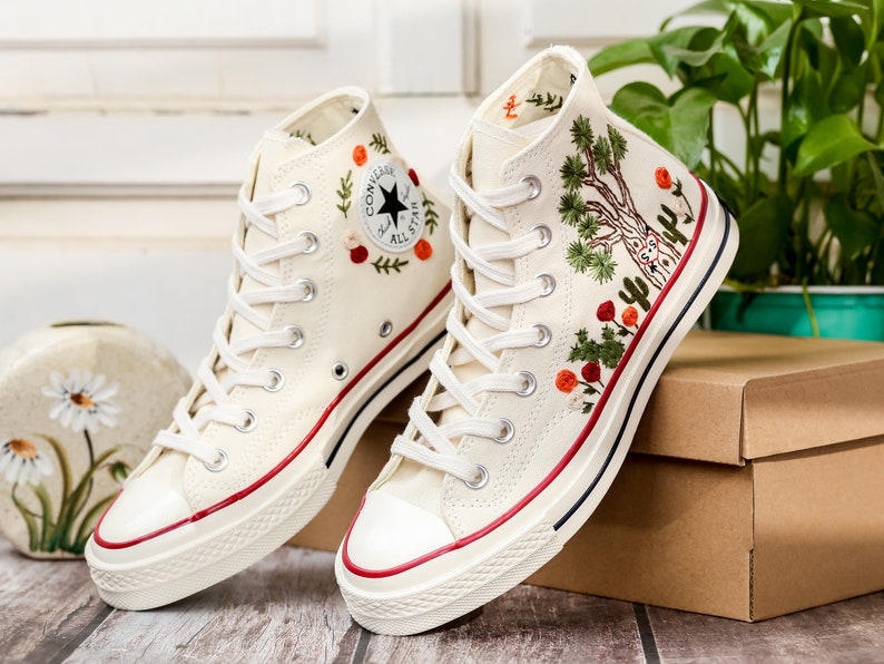 May include: A pair of white high-top canvas sneakers with red and green embroidered floral designs. The sneakers have a white rubber sole and a red stripe around the top.