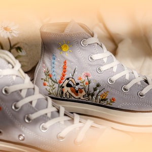 May include: A pair of gray high-top canvas sneakers with white laces. The sneakers are embroidered with a cat, flowers, and a sun.