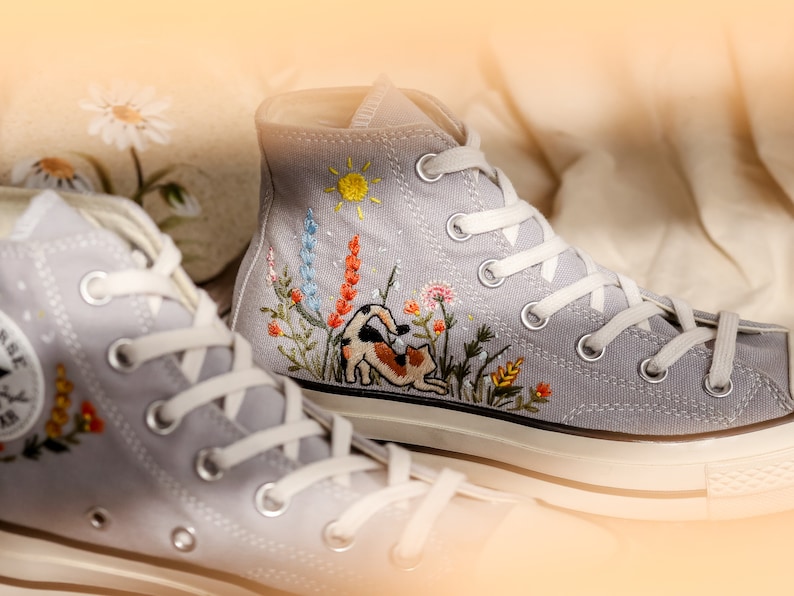 May include: A pair of gray high-top canvas sneakers with white laces. The sneakers are embroidered with a cat, flowers, and a sun.