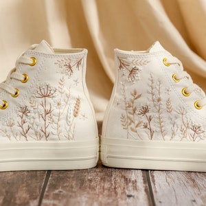 May include: A pair of white canvas high-top trainers with delicate floral embroidery in shades of brown and white.