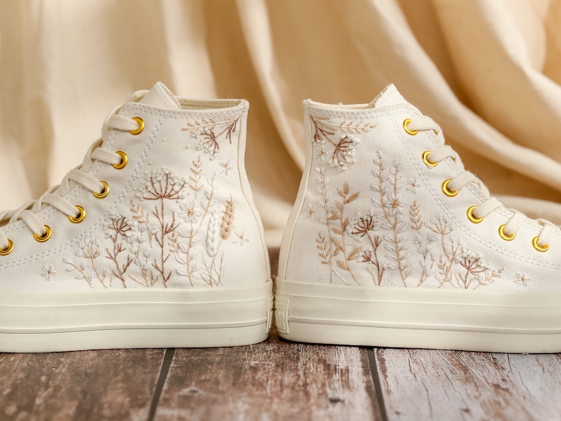 May include: A pair of white canvas high-top trainers with delicate floral embroidery in shades of brown and white.