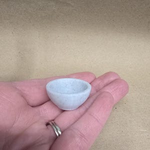 May include: A small, light blue speckled bowl. The bowl is held in a person's hand. The bowl is round with a slightly flared rim. The bowl is made of a speckled material.