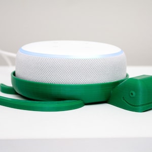 May include: A white smart speaker with a green 3D printed turtle stand. The turtle has a smiling face and a textured shell.