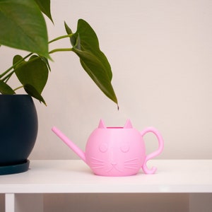 May include: A pink cat-shaped watering can with a spout and a tail. The watering can is sitting on a white shelf with a green plant in a dark blue pot.