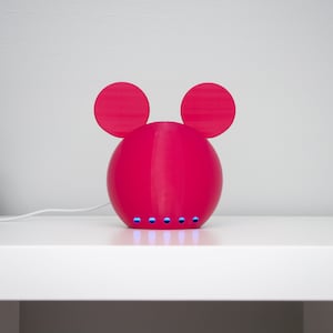 May include: A bright pink, spherical decorative object with two circular ears on top and a series of blue lights at the bottom. The object is on a white surface, with a small plant in a decorative pot to the left.