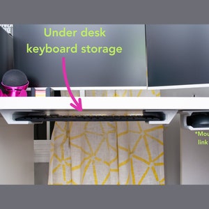 May include: White desk with a black keyboard stored underneath. The keyboard is attached to a white metal bracket. A black mouse holder is attached to the right side of the desk. The text "Under desk keyboard storage" is written in green on a pink arrow pointing to the keyboard. The text "*Mouse holder link in listing" is written in green below the mouse holder.