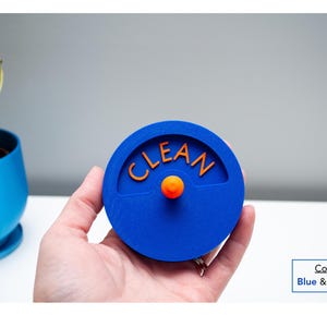 May include: A circular blue dishwasher magnet with the word CLEAN embossed in orange lettering. The center features a small orange knob that protrudes from the surface. The design is flat and round, providing a clear visual indicator for appliances.