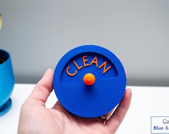 May include: A circular blue dishwasher magnet with the word CLEAN embossed in orange lettering. The center features a small orange knob that protrudes from the surface. The design is flat and round, providing a clear visual indicator for appliances.