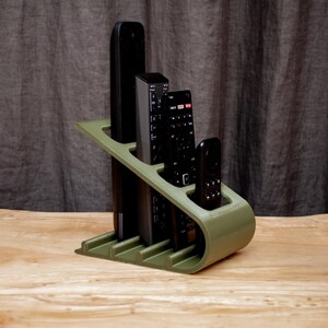 May include: A green plastic remote control organizer with four slots for remotes. The organizer is on a light wood surface. The remotes are black and gray.