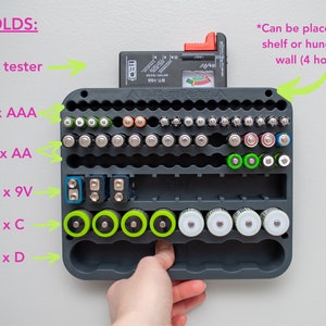 May include: A dark gray plastic battery organizer with designated slots for 38 AAA, 28 AA, 10 9V, 8 C, and 6 D batteries. The unit features a top slot for a .7-inch wide battery tester and includes four holes for wall mounting or shelf placement.
