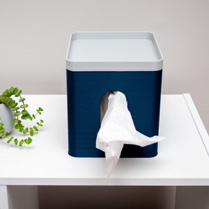 May include: A blue tissue box dispenser with a white top and a white tissue peeking out of the opening. The dispenser is sitting on a white shelf.