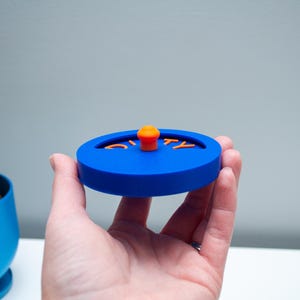 K&ouml;nnte beinhalten: Ein kreisf&ouml;rmiger blauer Deckel mit einem kleinen orangefarbenen Knopf in der Mitte und dem Wort DIRTY in orangefarbenen Buchstaben. Der Deckel hat einen vertieften halbkreisf&ouml;rmigen Ausschnitt, in dem der Text platziert ist, was ein funktionales Design ergibt.