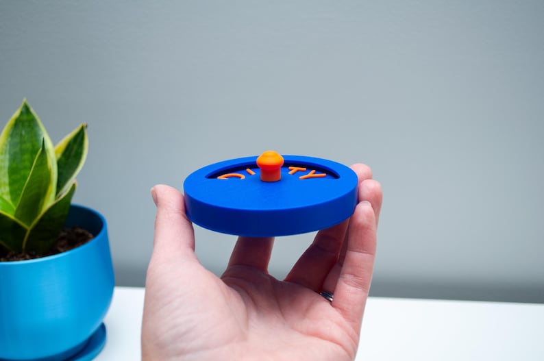 K&ouml;nnte beinhalten: Ein kreisf&ouml;rmiger blauer Deckel mit einem kleinen orangefarbenen Knopf in der Mitte und dem Wort DIRTY in orangefarbenen Buchstaben. Der Deckel hat einen vertieften halbkreisf&ouml;rmigen Ausschnitt, in dem der Text platziert ist, was ein funktionales Design ergibt.