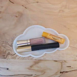 May include: Three makeup products, a lip gloss, mascara, and lip balm, are arranged on a white cloud-shaped tray. The lip gloss is a light pink color and has a gold cap. The mascara is black and has a gold cap. The lip balm is yellow and has a white label with black text. The text on the lip balm reads "Vitamin & Mineral Lip Treatment, SPF 15,  Free Speed,  Made in USA."
