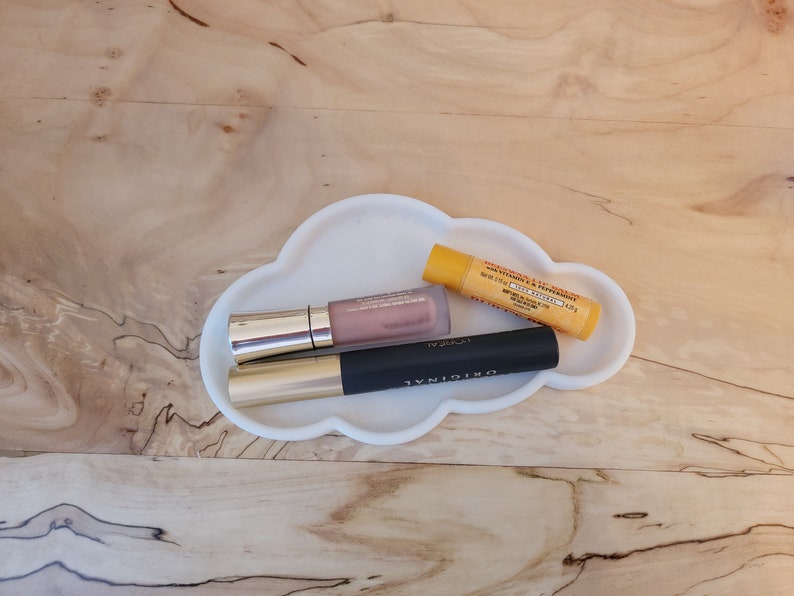 May include: Three makeup products, a lip gloss, mascara, and lip balm, are arranged on a white cloud-shaped tray. The lip gloss is a light pink color and has a gold cap. The mascara is black and has a gold cap. The lip balm is yellow and has a white label with black text. The text on the lip balm reads "Vitamin & Mineral Lip Treatment, SPF 15,  Free Speed,  Made in USA."