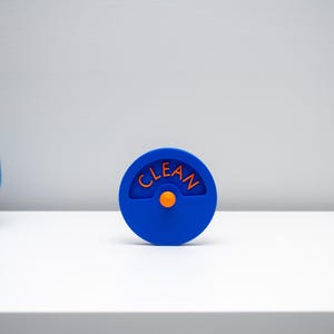 K&ouml;nnte beinhalten: Ein kreisf&ouml;rmiger blauer Sp&uuml;lmaschinenmagnet mit einem zentralen orangefarbenen Knopf und einem bogenf&ouml;rmigen Ausschnitt, der das Wort CLEAN in orangefarbenen Gro&szlig;buchstaben enth&auml;lt. Der Magnet hat eine glatte Oberfl&auml;che und ein schlichtes Design.