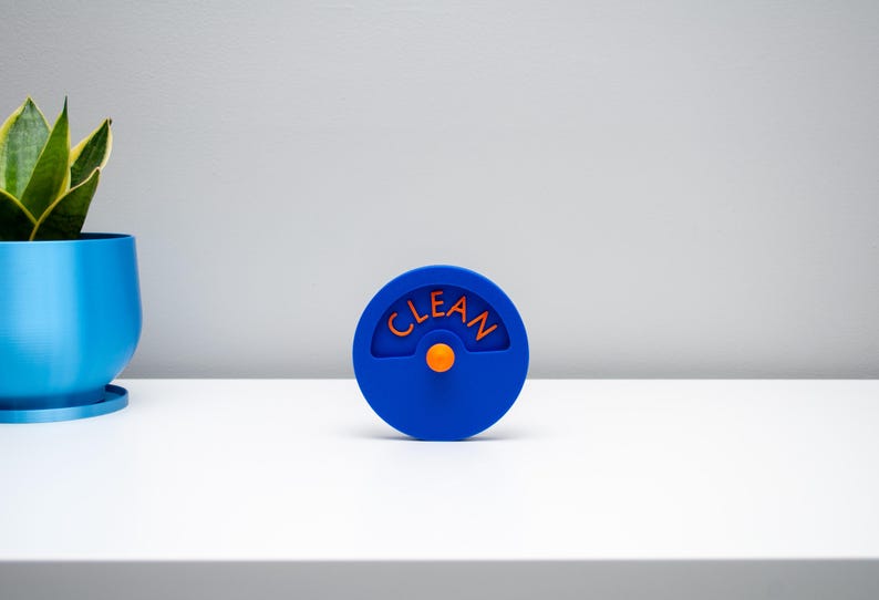 K&ouml;nnte beinhalten: Ein kreisf&ouml;rmiger blauer Sp&uuml;lmaschinenmagnet mit einem zentralen orangefarbenen Knopf und einem bogenf&ouml;rmigen Ausschnitt, der das Wort CLEAN in orangefarbenen Gro&szlig;buchstaben enth&auml;lt. Der Magnet hat eine glatte Oberfl&auml;che und ein schlichtes Design.