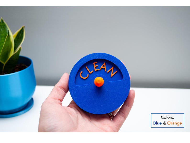 K&ouml;nnte beinhalten: Ein kreisf&ouml;rmiger blauer Sp&uuml;lmaschinenmagnet mit dem Wort CLEAN in orangefarbenen Buchstaben. In der Mitte befindet sich ein kleiner orangefarbener Knopf, der aus der Oberfl&auml;che herausragt. Das Design ist flach und rund und dient als Anzeige.