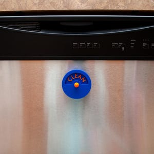 K&ouml;nnte beinhalten: Ein kreisf&ouml;rmiger blauer Sp&uuml;lmaschinenmagnet mit dem Wort CLEAN in orangefarbenen Buchstaben. Der Magnet verf&uuml;gt &uuml;ber einen kleinen orangefarbenen Knopf in der Mitte und ist an der Vorderseite einer Edelstahl-Sp&uuml;lmaschine mit schwarzem Bedienfeld.