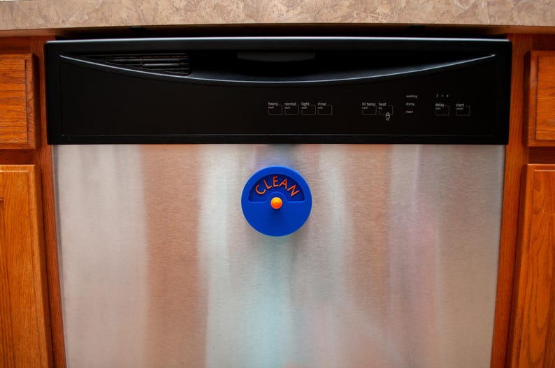 K&ouml;nnte beinhalten: Ein kreisf&ouml;rmiger blauer Sp&uuml;lmaschinenmagnet mit dem Wort CLEAN in orangefarbenen Buchstaben. Der Magnet verf&uuml;gt &uuml;ber einen kleinen orangefarbenen Knopf in der Mitte und ist an der Vorderseite einer Edelstahl-Sp&uuml;lmaschine mit schwarzem Bedienfeld.