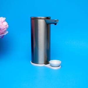 May include: A stainless steel soap dispenser with a small, attached dish, set against a bright blue background. The dispenser has a cylindrical body and a spout. A bouquet of pink peonies is visible on the left.