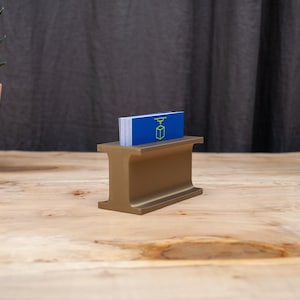 May include: A gold-colored business card holder in the shape of an I-beam, holding a stack of blue business cards. The cards have a green logo. The holder sits on a wooden surface, with a potted plant in the background.