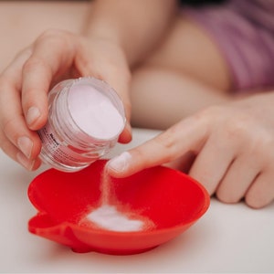 May include: A clear jar of pink powder is being poured onto a finger and into a red, heart-shaped dish. The powder is a light pink color. The dish is made of a flexible material.