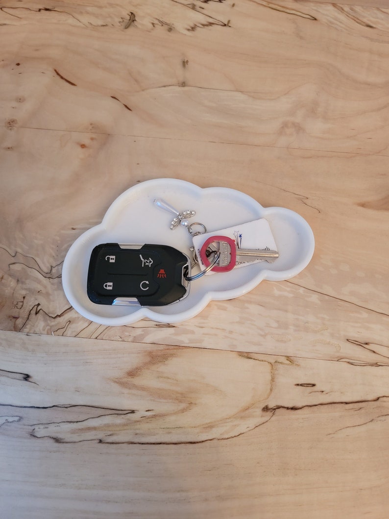 May include: White ceramic cloud-shaped dish with a car key and a key chain with a small charm. The key chain is attached to a key with a red plastic tag.