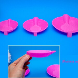 May include: Three fuchsia-colored, oval-shaped trays with attached spoon-like handles. The trays are arranged from smallest to largest, with a rectangular base supporting each. The background is a bright blue.