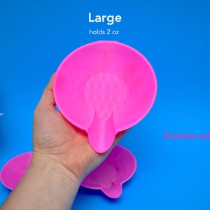 May include: A fuchsia-colored, large, round dish with a small handle. The dish holds 2 oz. The text "Large" and "holds 2 oz" are above the dish. The text "[Fuchsia color]" is below the dish. The dish is held by a hand against a blue background.