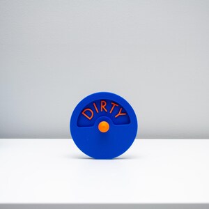 K&ouml;nnte beinhalten: Ein kreisf&ouml;rmiger blauer Sp&uuml;lmaschinenmagnet mit einem drehbaren Mittelrad. Das Wort DIRTY ist in orangefarbenen Buchstaben auf der oberen H&auml;lfte des Kreises eingepr&auml;gt, und ein kleiner orangefarbener Knopf dient zum Drehen der Anzeige.
