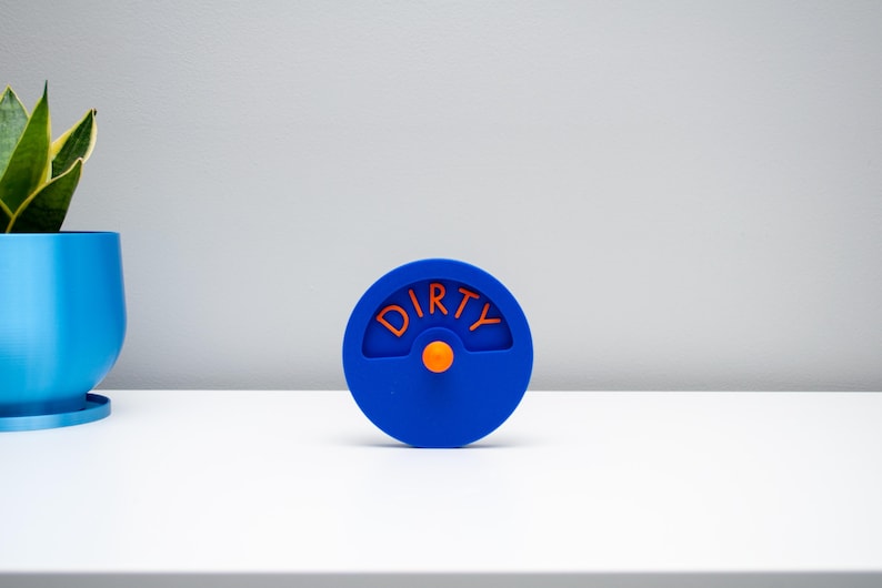K&ouml;nnte beinhalten: Ein kreisf&ouml;rmiger blauer Sp&uuml;lmaschinenmagnet mit einem drehbaren Mittelrad. Das Wort DIRTY ist in orangefarbenen Buchstaben auf der oberen H&auml;lfte des Kreises eingepr&auml;gt, und ein kleiner orangefarbener Knopf dient zum Drehen der Anzeige.