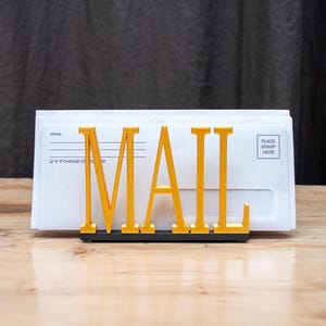 May include: A gold metal mail holder with the word "MAIL" in large, bold letters. The holder is sitting on a wooden surface and has a white envelope in it.