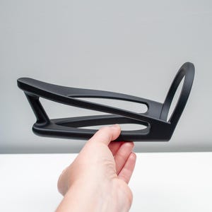 May include: A matte black bicycle water bottle cage featuring a sleek, open-frame design with a curved base and a circular top support. The structure is composed of smooth, continuous lines that form a lightweight, aerodynamic shape for holding bottles.