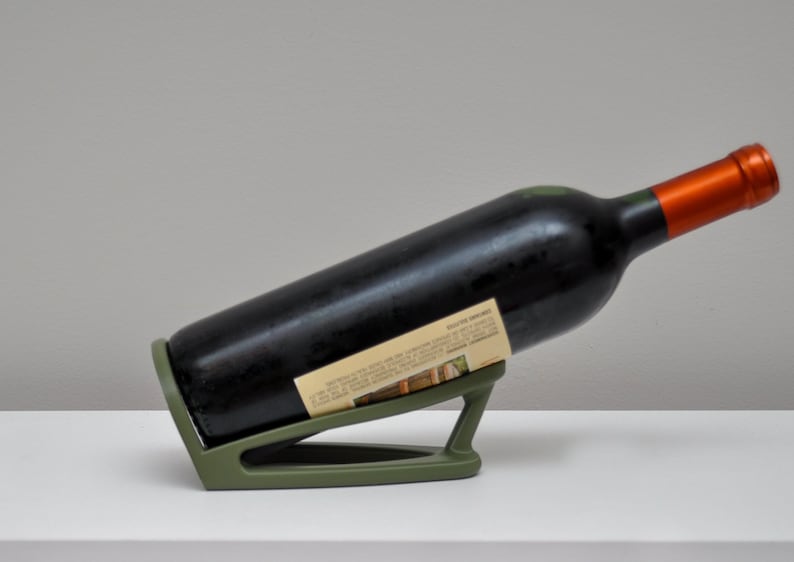 K&ouml;nnte beinhalten: Ein gr&uuml;ner Flaschenhalter aus Kunststoff mit einer Flasche Rotwein, die darauf liegt. Die Flasche hat ein Etikett mit Text und einem Bild eines Weinbergs.