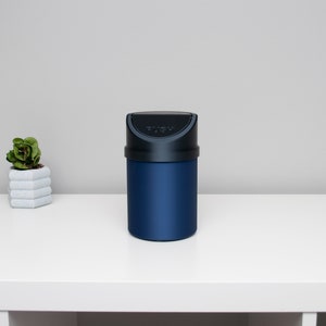 May include: A blue trash can with a black lid and the word "PUSH" printed on the lid. The trash can is sitting on a white surface.