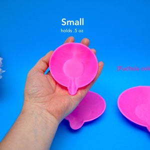 May include: Bright fuchsia-colored small dish with a unique shape, holding 0.5 oz. The dish is held in a hand against a vibrant blue background. The text "Small holds .5 oz" is visible.