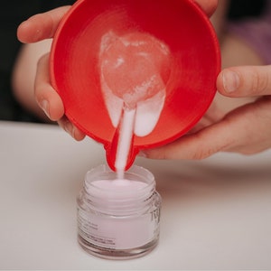 May include: A red bowl is pouring pink powder into a small glass jar. The jar is filled with pink powder. The image is taken from a close-up angle, with the hands holding the bowl and jar in focus.