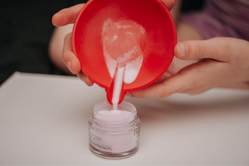 May include: A red bowl is pouring pink powder into a small glass jar. The jar is filled with pink powder. The image is taken from a close-up angle, with the hands holding the bowl and jar in focus.
