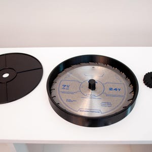 May include: A circular saw blade with a black plastic housing, a black gear, and a black disc with a central hole. The saw blade is silver with blue text, and the housing is black. The blade is 7 inches in diameter.