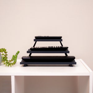 May include: A tiered, dark gray remote control organizer. The organizer has three levels, each holding a black remote. The design is minimalist and modern, suitable for home entertainment systems.