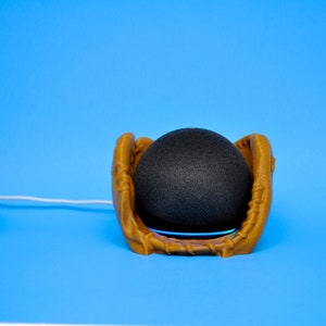 May include: A black smart speaker sits inside a tan baseball glove-shaped holder, with a blue light emanating from the base. A small plant in a silver holder is to the left. The background is a solid blue.