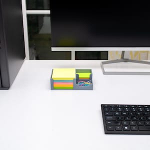 May include: A gray desk organizer containing a stack of square yellow sticky notes and a smaller compartment holding a stack of smaller yellow sticky notes alongside a collection of colorful metal paper clips in blue, pink, and green.
