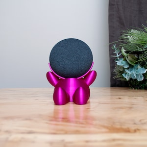 May include: A magenta-colored, 3D-printed stand cradles a dark gray smart speaker. The stand has a stylized, rounded shape with arms that curve up to hold the speaker. The speaker is a small, spherical device. The stand is on a wooden surface.